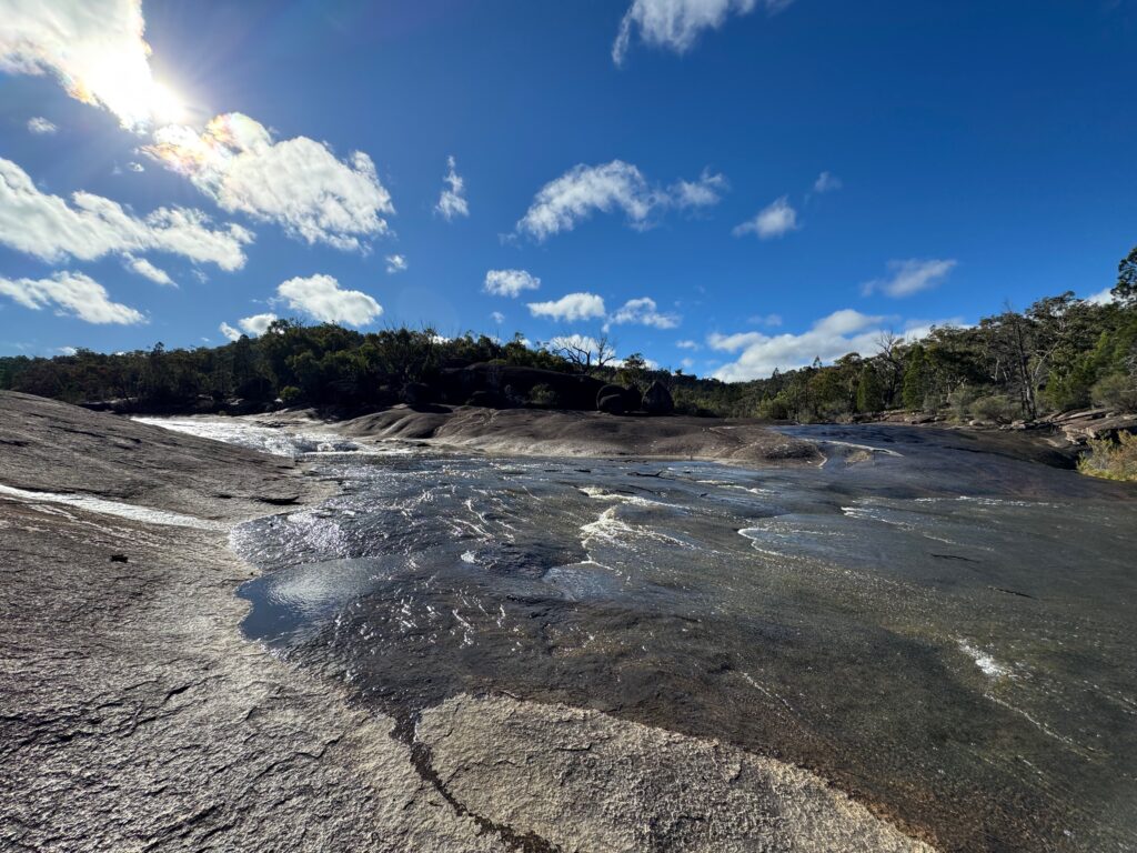 Water moving across a broad granite slab under a blue sky with light cloud